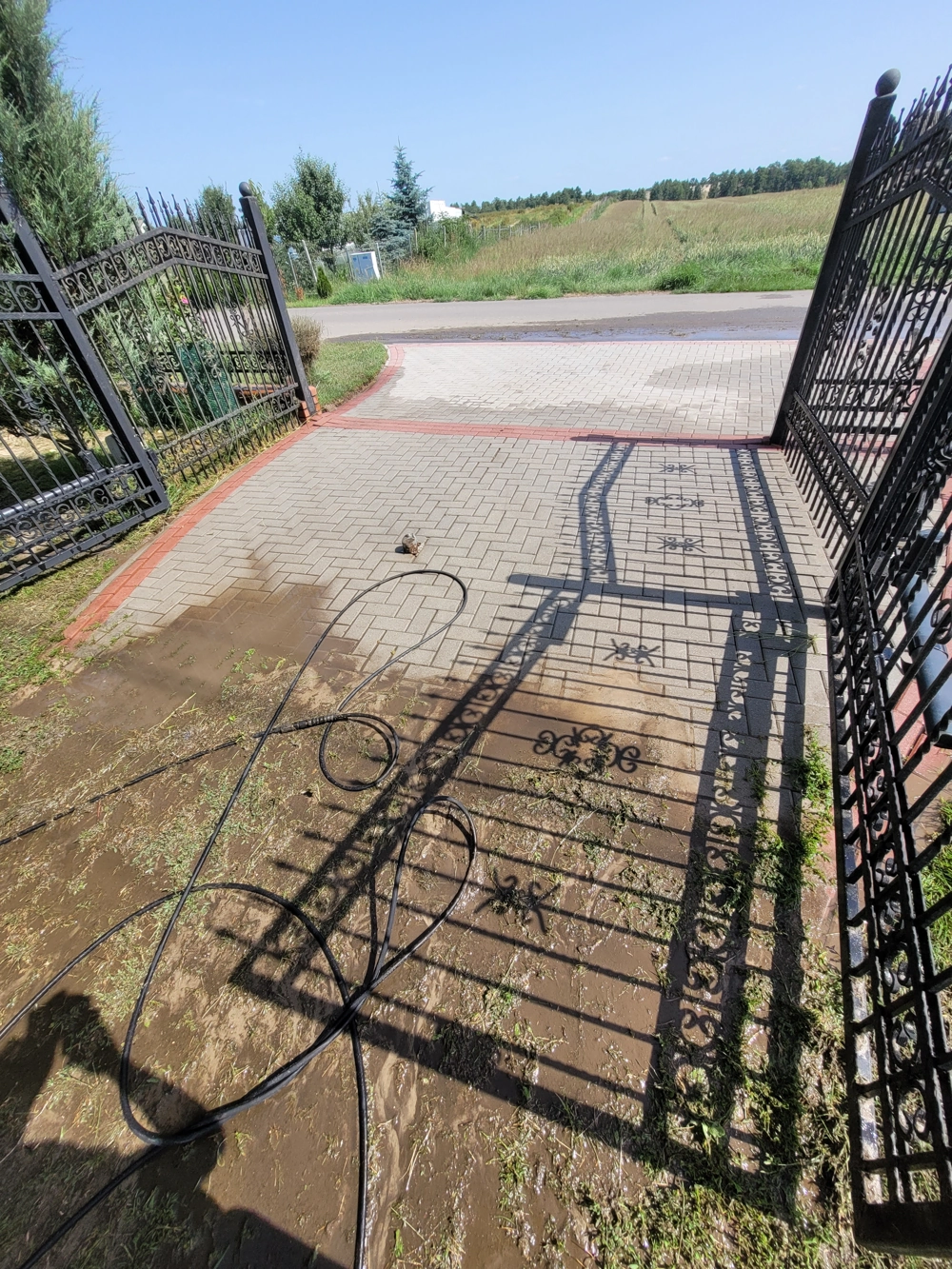 Wąż ciśnieniowy na mokrej kostce brukowej przy otwartej kutej bramie – mycie kostki brukowej